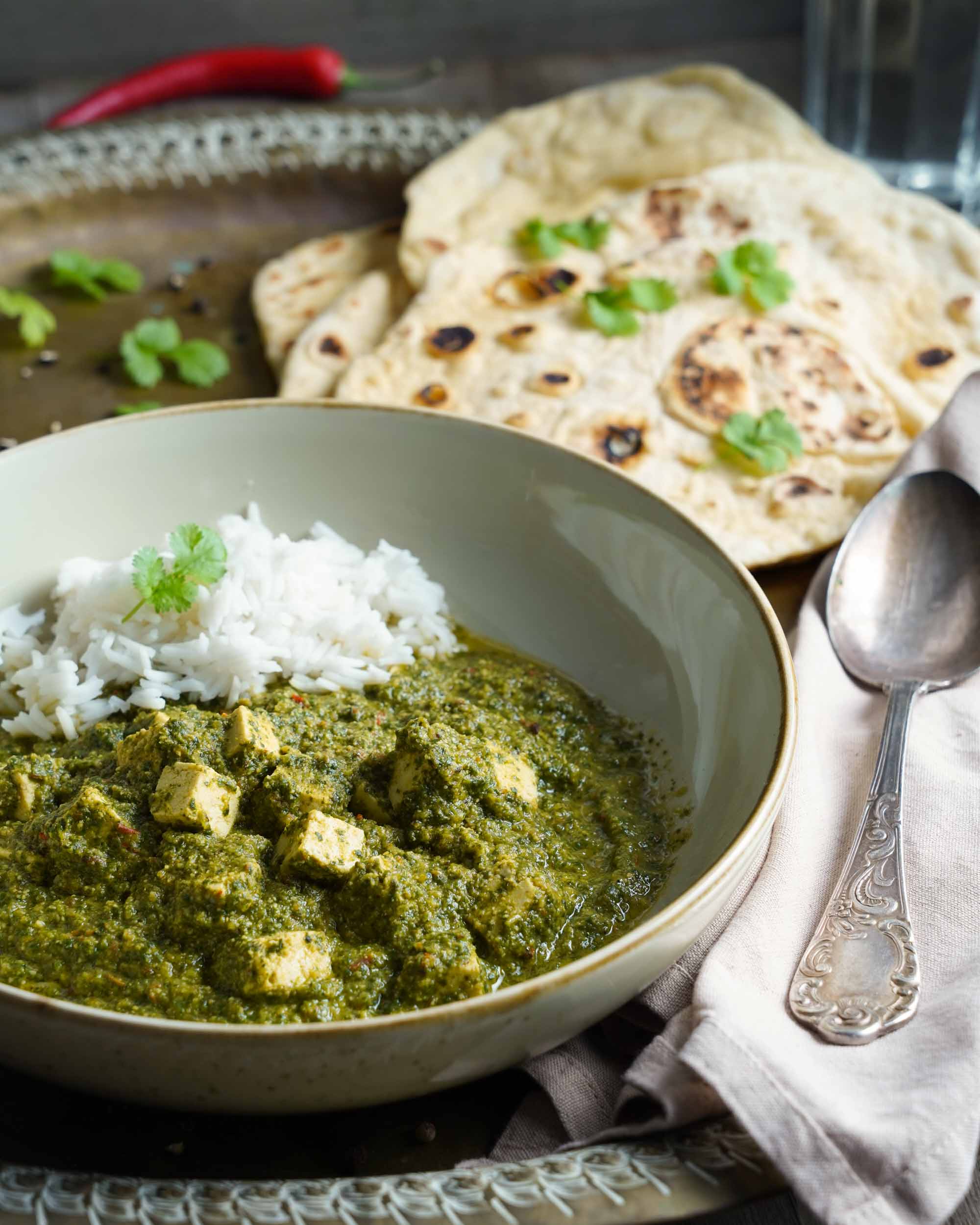 Vegane Palak Paneer in zwei Schüsseln, dazu Naanbrot, Wasser in perspektivischer Ansicht im Detail. Dekoriert mit frischen Zutaten wie Chili und Knoblauch.