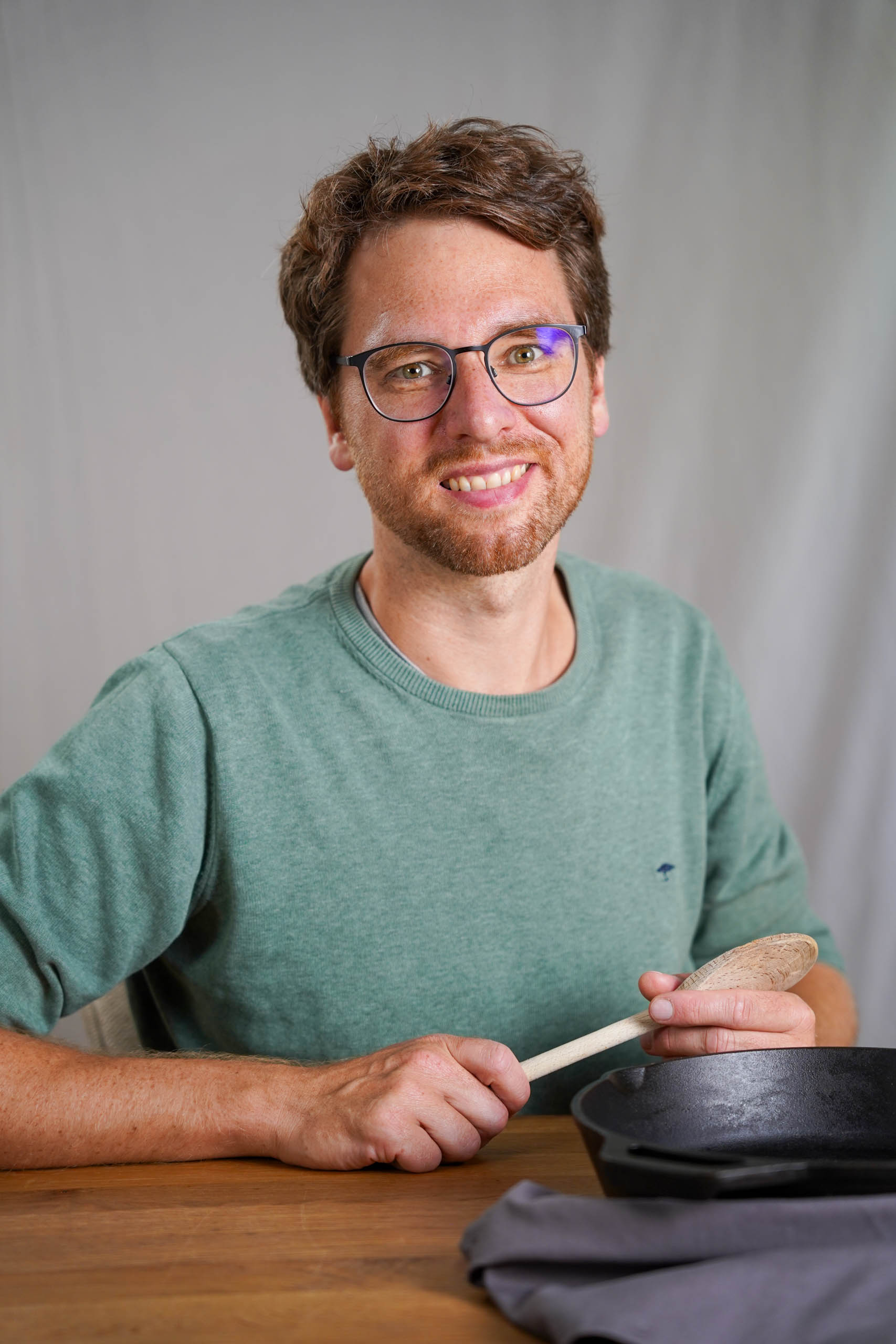 Portrait, wo ich an einem Holztisch sitze. Kochlöffel in der Hand und gußeiserne Pfanne vor mir.